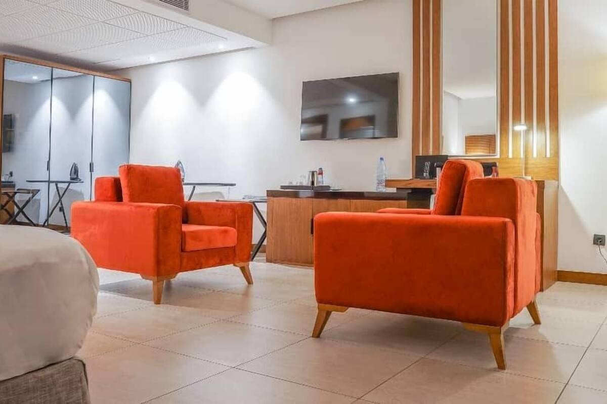 A hotel room with a bed, two orange armchairs, a small table, a desk, and a mirror.