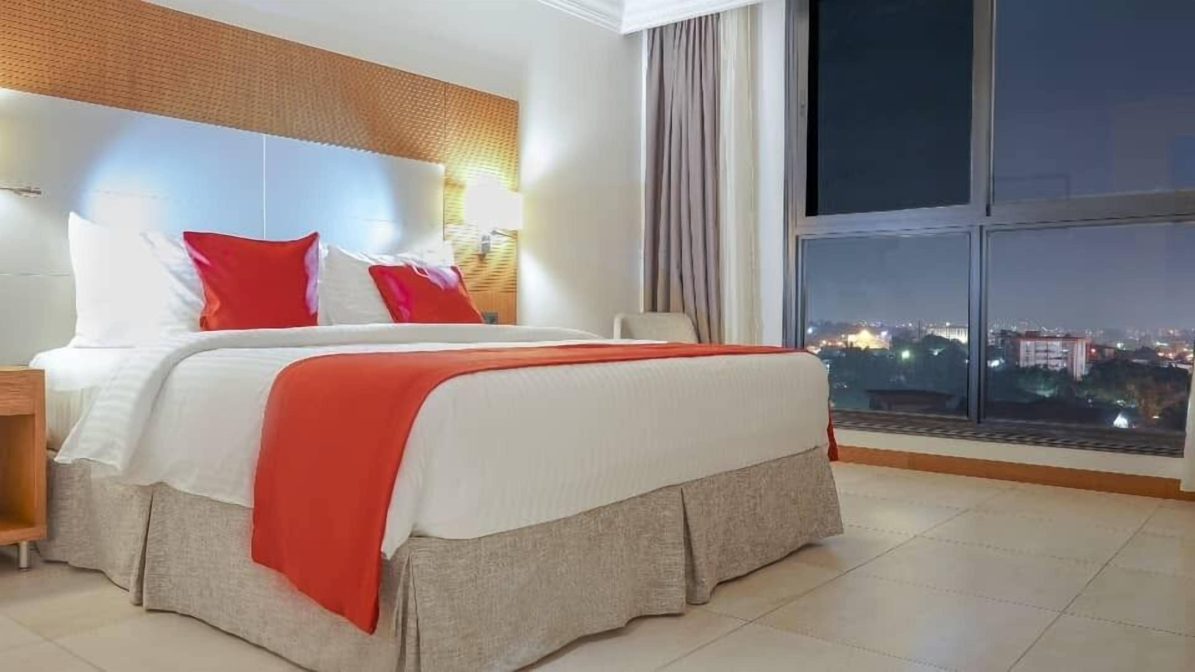 A modern hotel room with a large bed, a nightstand, a window with a city view, and a wooden headboard.