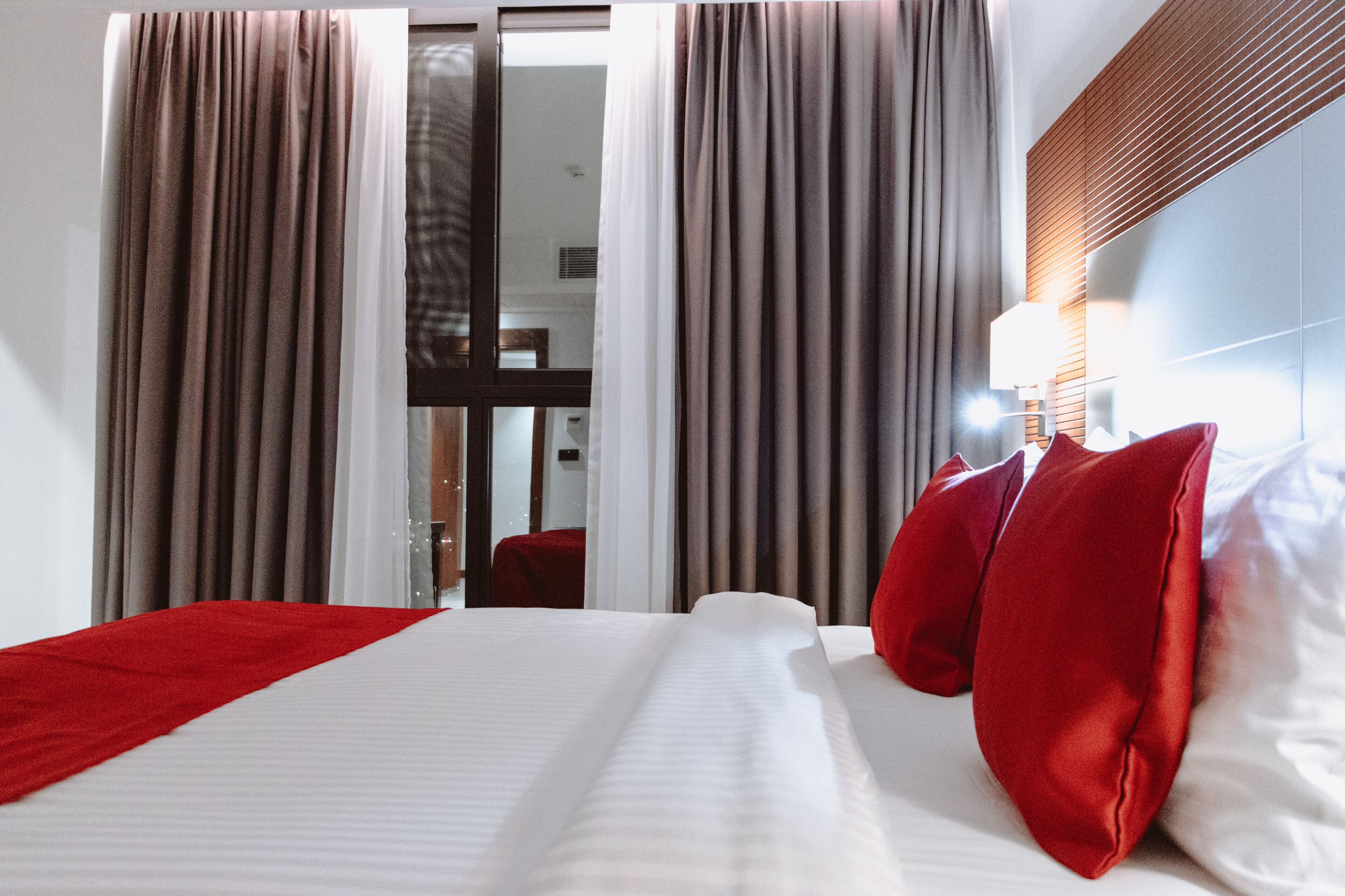 Quarto de hotel com uma cama grande, almofadas vermelhas e uma janela com cortinas.