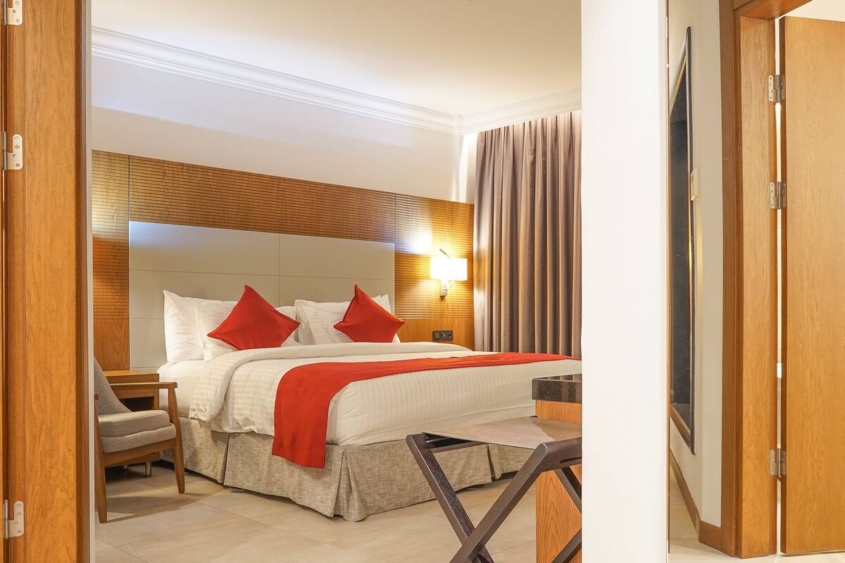 A hotel room with a bed, a nightstand, a chair, a desk, and a wooden headboard.
