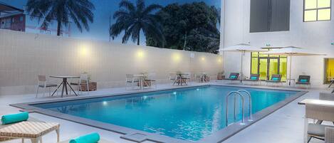 A swimming pool with lounge chairs and tables, surrounded by a tiled wall and palm trees.