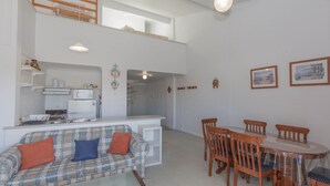A spacious living area with a sofa, dining table, and chairs. A kitchen with a microwave and stove is visible in the background. - Razorback 1/1 Gippsland St (Jindabyne)
