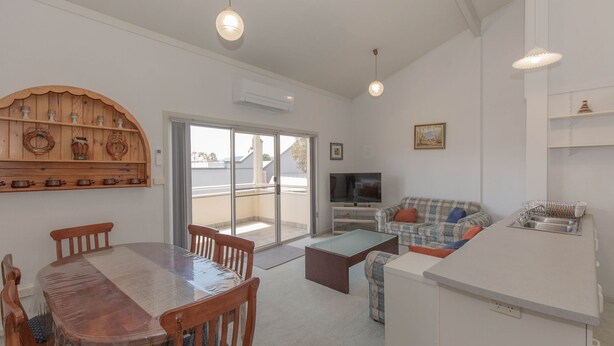 A modern living room with a dining table, chairs, a sofa, a TV, and a kitchen area with a sink and dish rack. - Razorback 1/1 Gippsland St (Jindabyne)