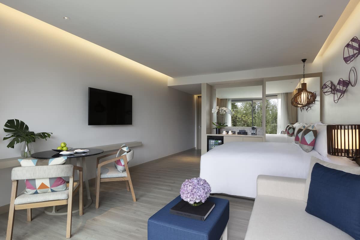 Una habitación de hotel moderna con una cama grande, un área de comedor con sillas, un televisor montado en la pared y vistas al exterior a través de amplios ventanales.