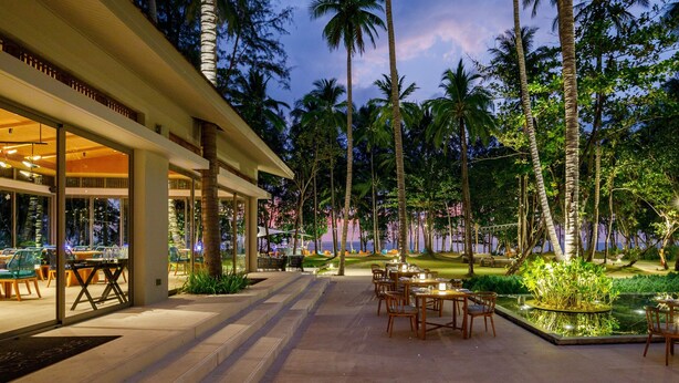 An outdoor dining area with tables and chairs, surrounded by palm trees and a pool.
