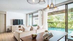 A modern living room with a wooden dining table, white sofa, and a TV. There is a pool visible through the large windows.