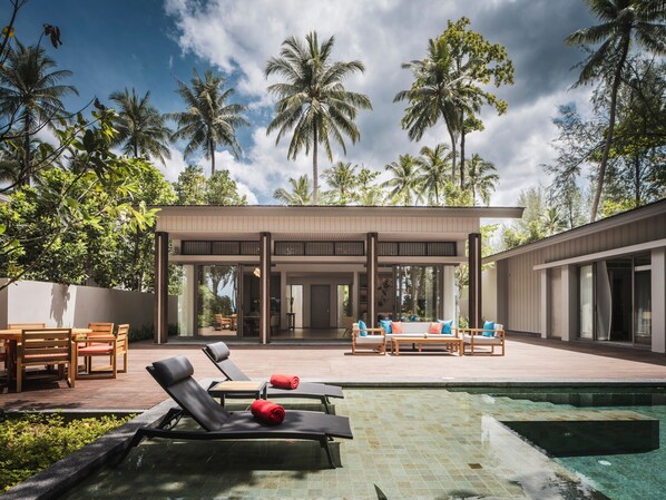 Un área de piscina moderna al aire libre con sillones de descanso y una zona de estar techada, rodeada de altas palmeras.