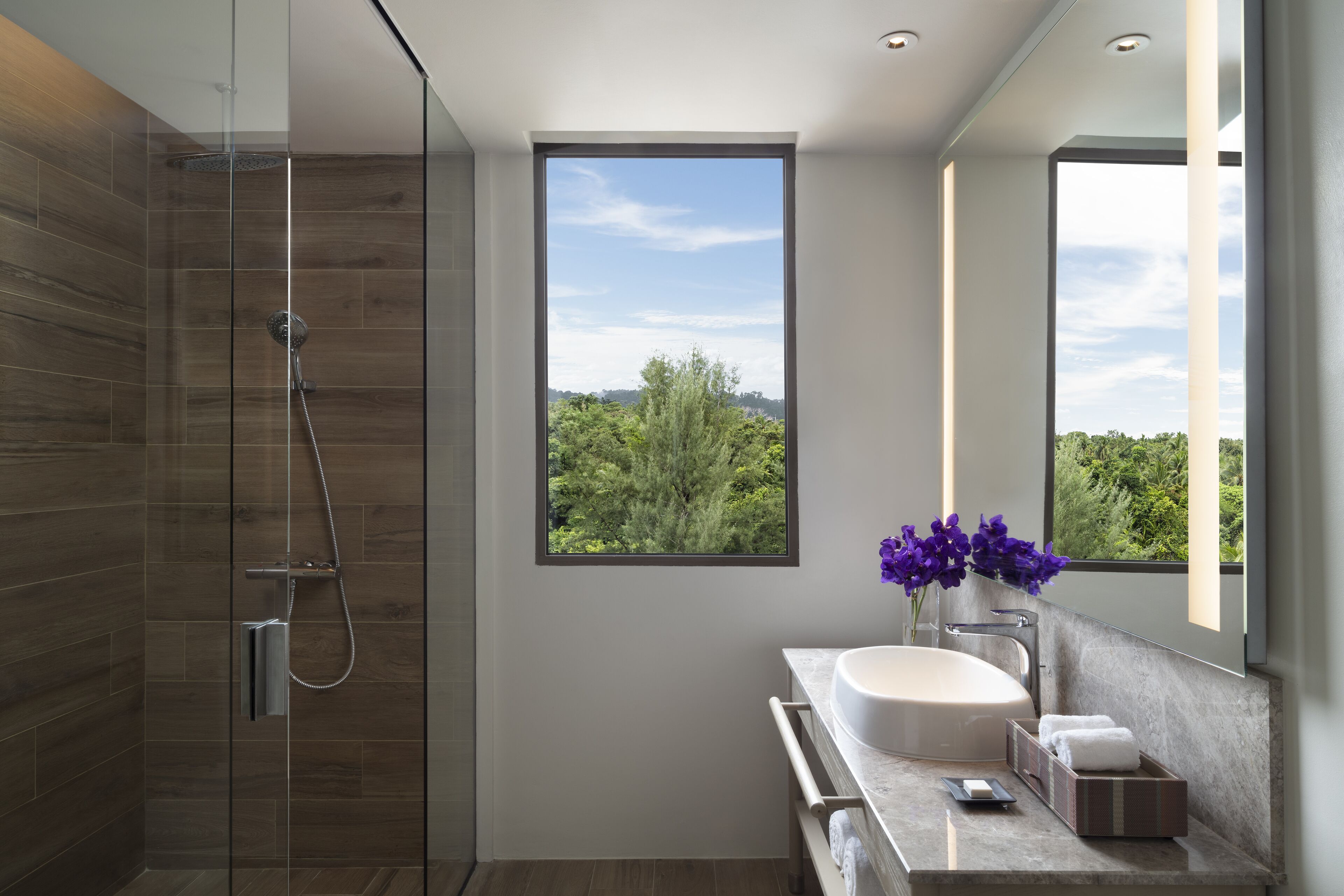 A modern bathroom with a glass-enclosed shower, a large window with a view of greenery, and a marble sink with a vase of purple flowers.