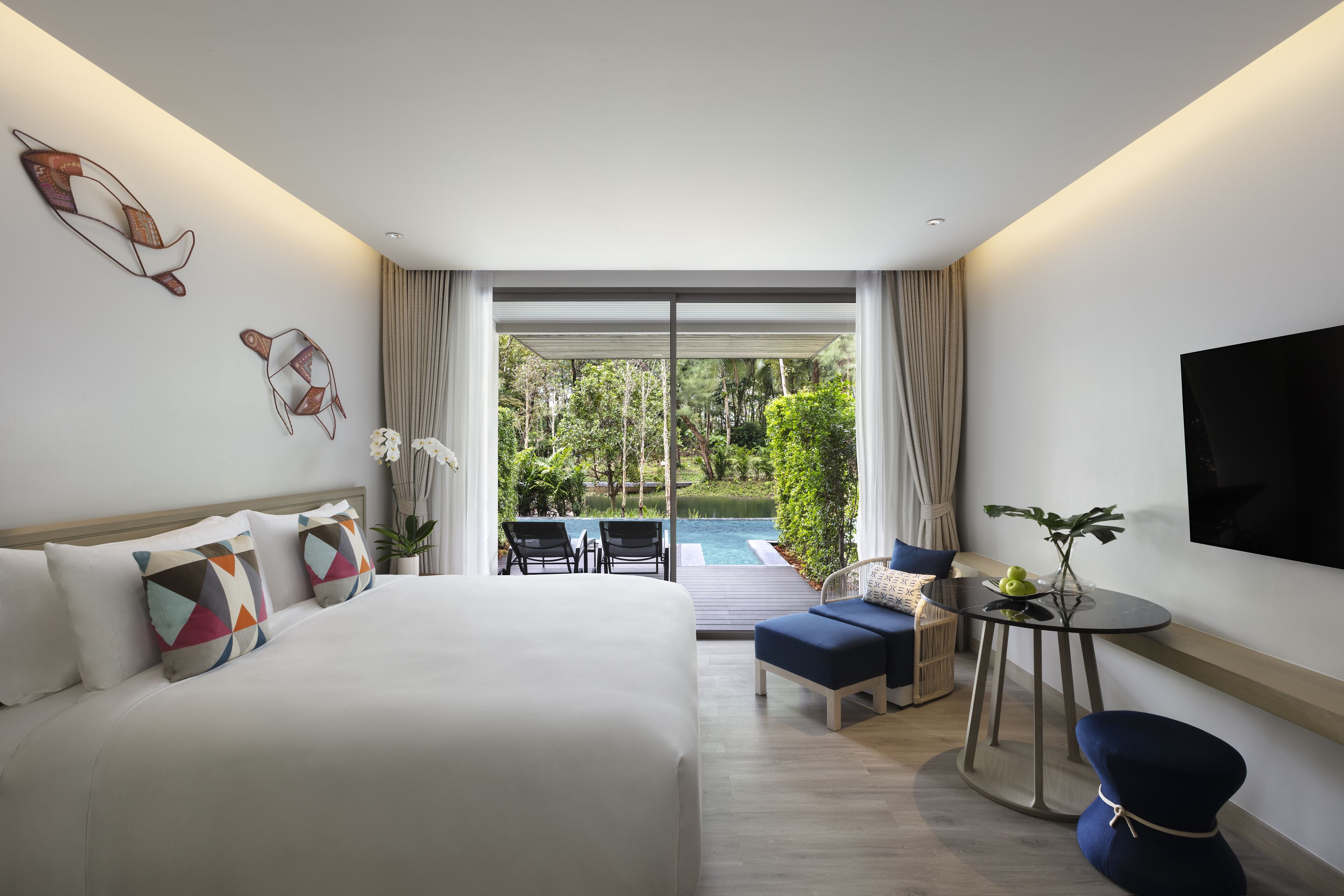 A modern hotel room with a large bed, a small table with a plant, a TV mounted on the wall, and a view of a pool and garden through large windows.