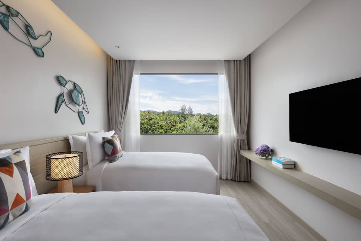 Una habitación de hotel moderna con dos camas, un televisor, una ventana con vistas a zonas verdes y una estantería con un libro y un jarrón.