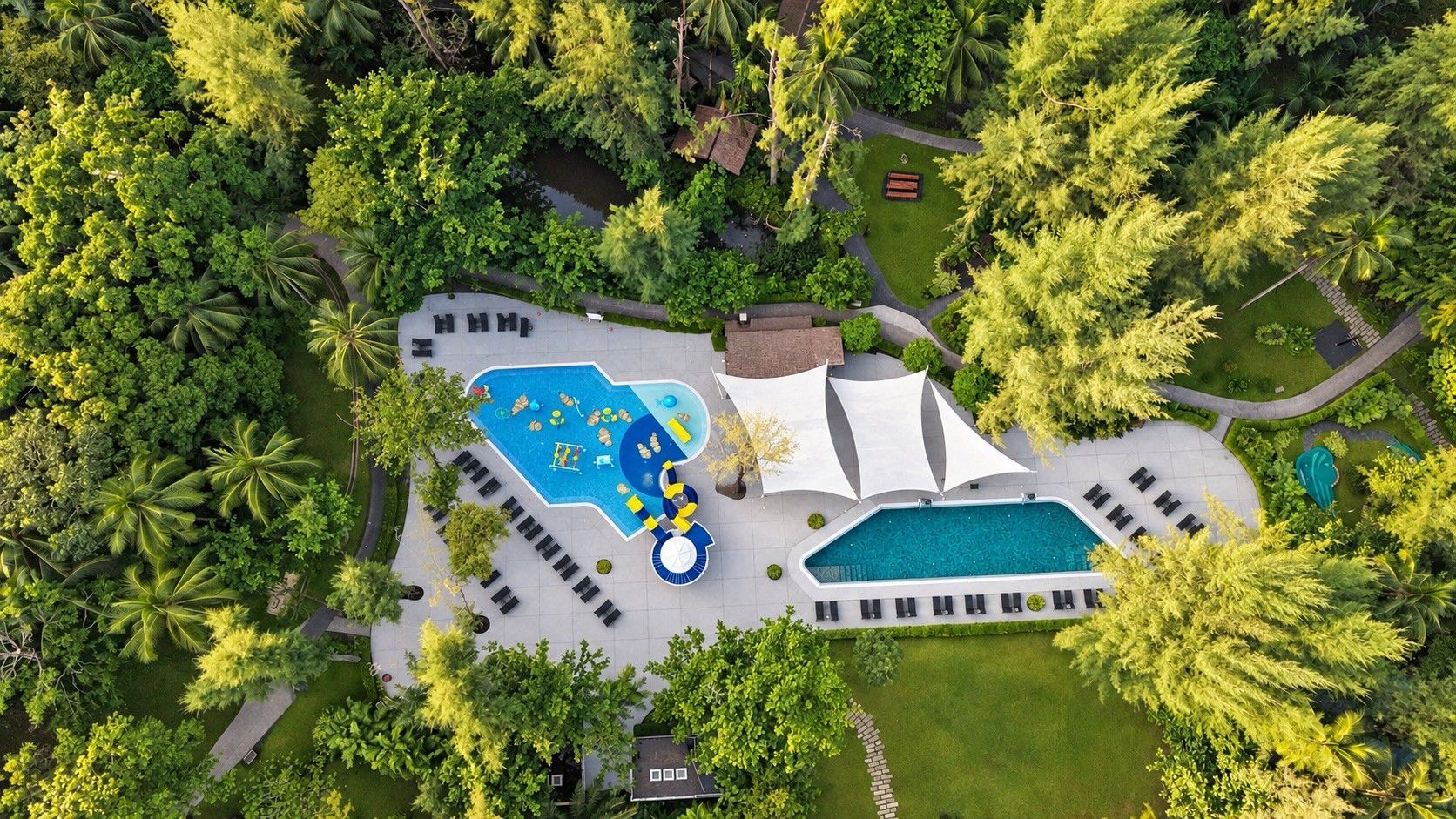 Vista aérea de un complejo turístico con una piscina grande, sillones de sol y un pabellón.