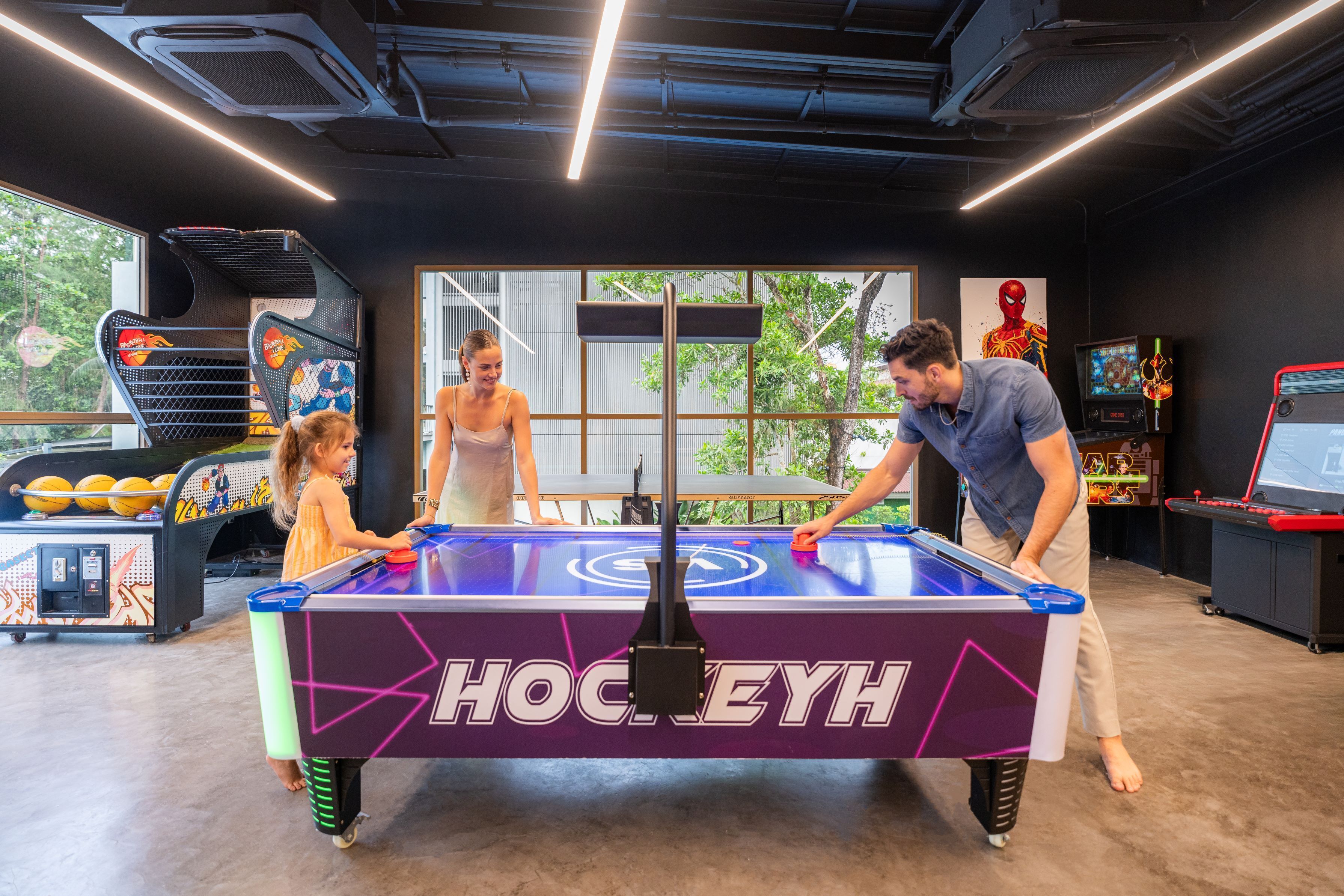 A table hockey game with a "HockeyH" logo, surrounded by arcade games and a large window with a view of trees.