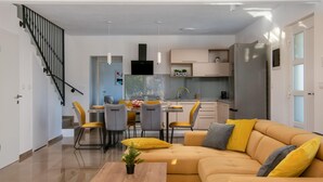 Een moderne woonkamer met een bank, een eettafel en stoelen. Er is een keuken met kastjes en een koelkast. Een trap is links te zien.