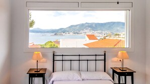 A bedroom with a large window overlooking a coastal town and mountains. There are two bedside tables with lamps. - Cap Ras 01 – Apartment with terrace and sea views (Llança)