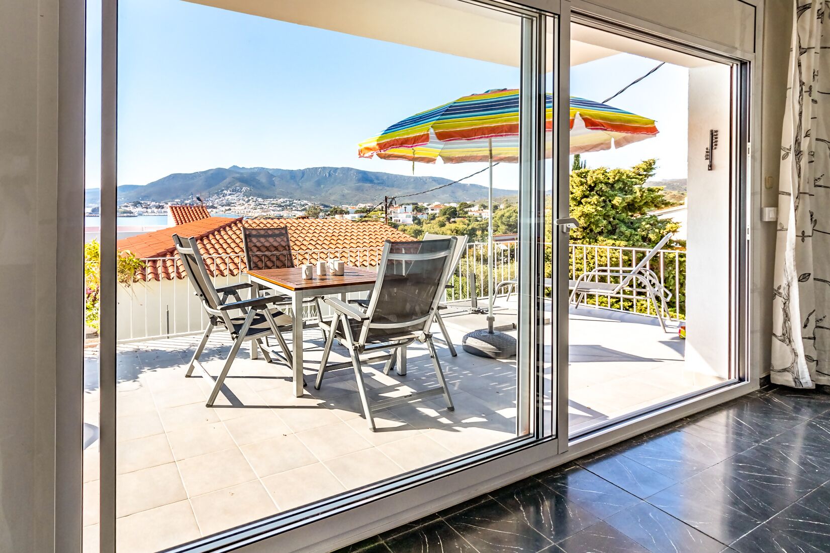 Een balkon met een tafel en stoelen, een kleurrijke parasol en uitzicht op een stad en bergen.