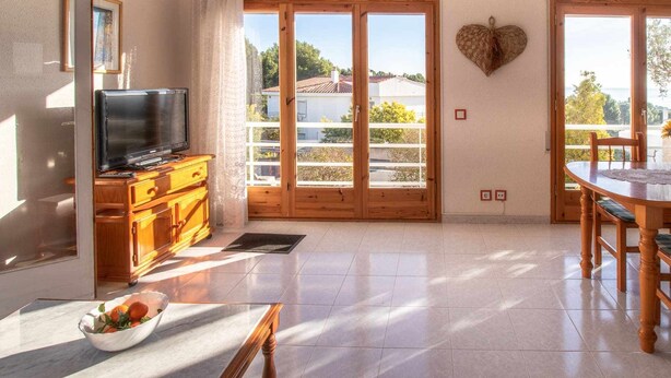 A room with a television on a wooden cabinet, a dining table with chairs, and a heart-shaped decoration on the wall. - Cap Ras 04 Comfortable apartment close to the sea (Llança)