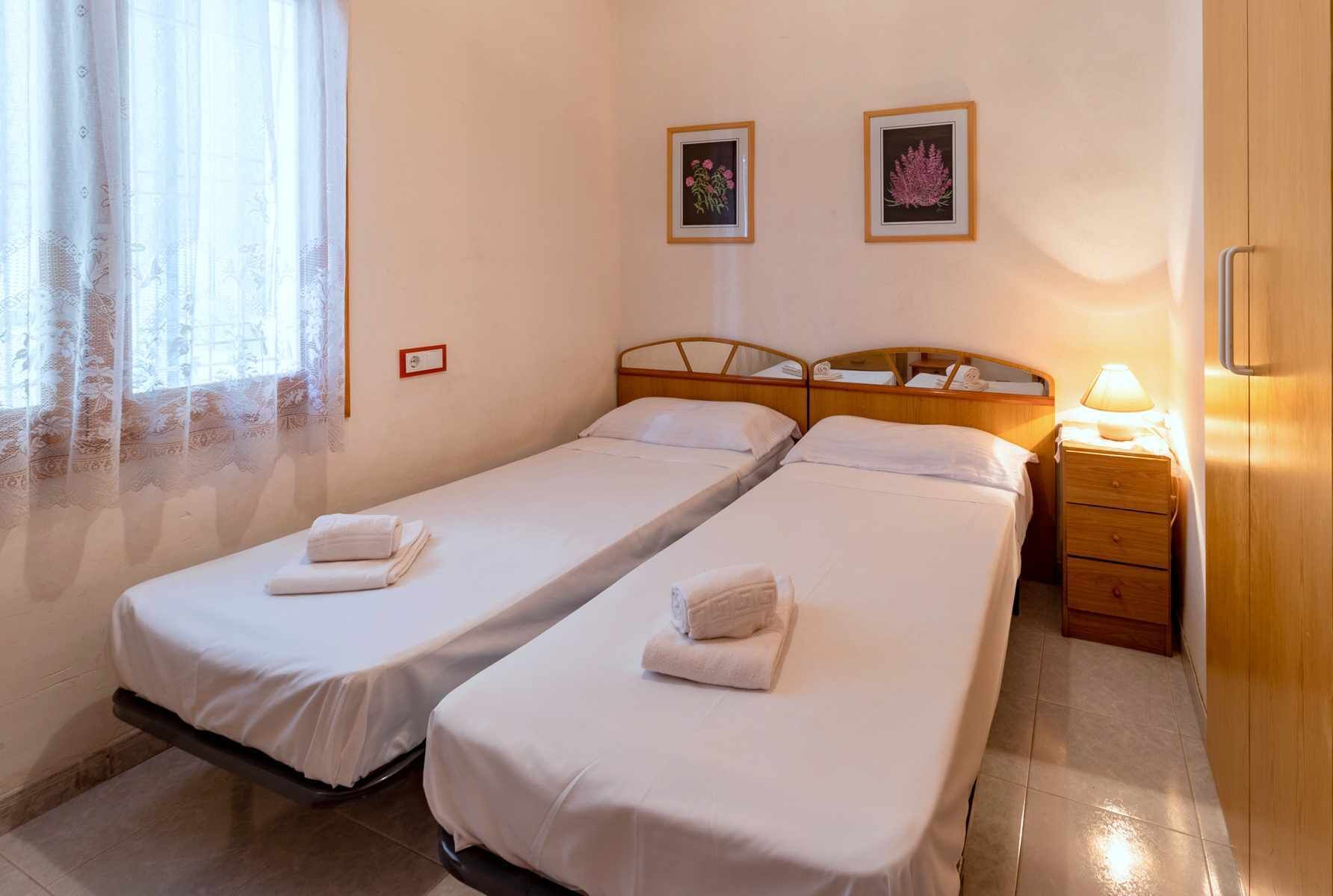 A hotel room with two single beds, a wooden nightstand with a lamp, and two framed pictures on the wall.