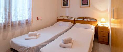 A hotel room with two single beds, a wooden nightstand with a lamp, and two framed pictures on the wall.