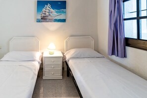 Habitación de hotel con dos camas individuales, una mesita de noche con lámpara, un cuadro de un velero y una ventana con cortinas.