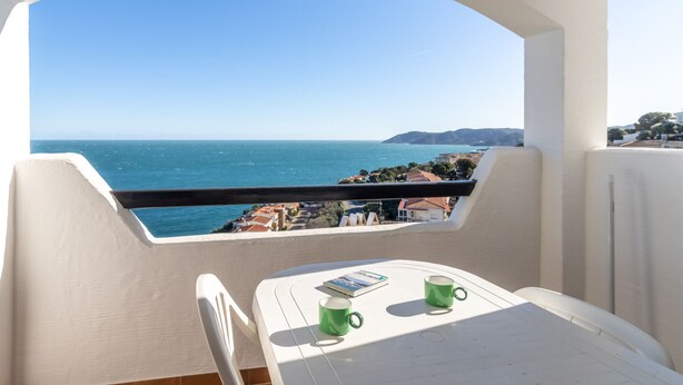 A balcony with a view of the sea, a table with two green cups, and a book.