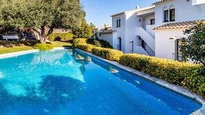 A swimming pool surrounded by white buildings with tiled roofs, a tree, and well-maintained hedges.