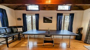 Una stanza con tavolo da ping pong, pavimento in legno, due finestre con tende, una panca e un quadro appeso al muro.