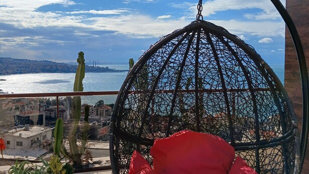 Un fauteuil suspendu avec des coussins rouges, placé sur un balcon donnant sur un paysage urbain et la mer.