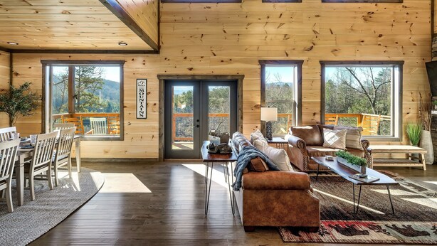 A spacious wooden interior with a dining area, seating, and large windows offering views of the outdoors. - Heated Large Indoor POOL, VIEWS, Firepit, Location! 6BRCabin-sleeps 20 Game Room (Sevierville)