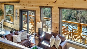 A spacious wooden cabin with multiple large windows, a cozy living area with a brown leather sofa, and a hanging light fixture. - Heated Large Indoor POOL, VIEWS, Firepit, Location! 6BRCabin-sleeps 20 Game Room (Sevierville)