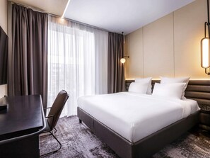 Ein Hotelzimmer mit einem Bett, einem Schreibtisch, einem Sessel und einem großen Fenster mit Vorhängen.