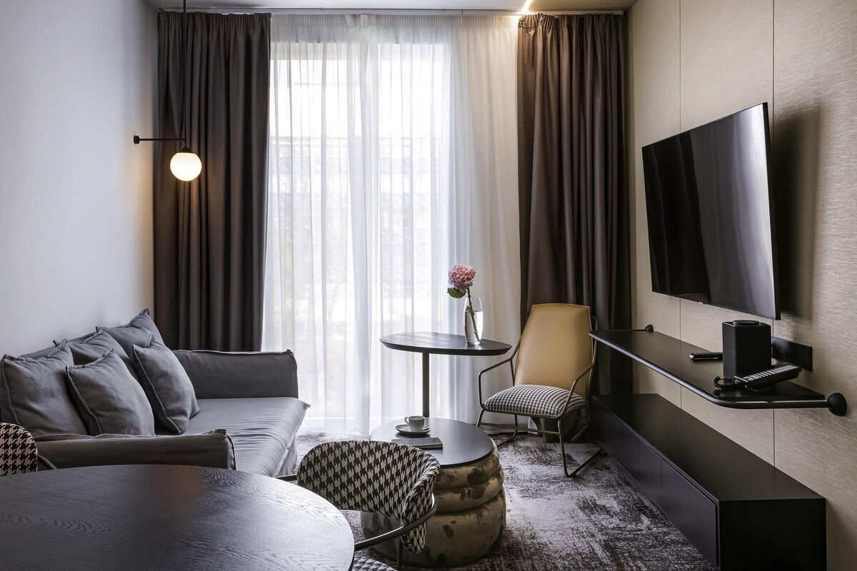 Une chambre d’hôtel moderne avec un canapé, une table basse ronde, une petite table d’appoint avec un vase, une chaise, un téléviseur fixé au mur et une grande fenêtre avec des rideaux.