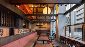 Un intérieur de restaurant moderne avec des tables et des bancs en bois, un plafond haut avec des poutres apparentes et de grandes fenêtres donnant sur la rue.