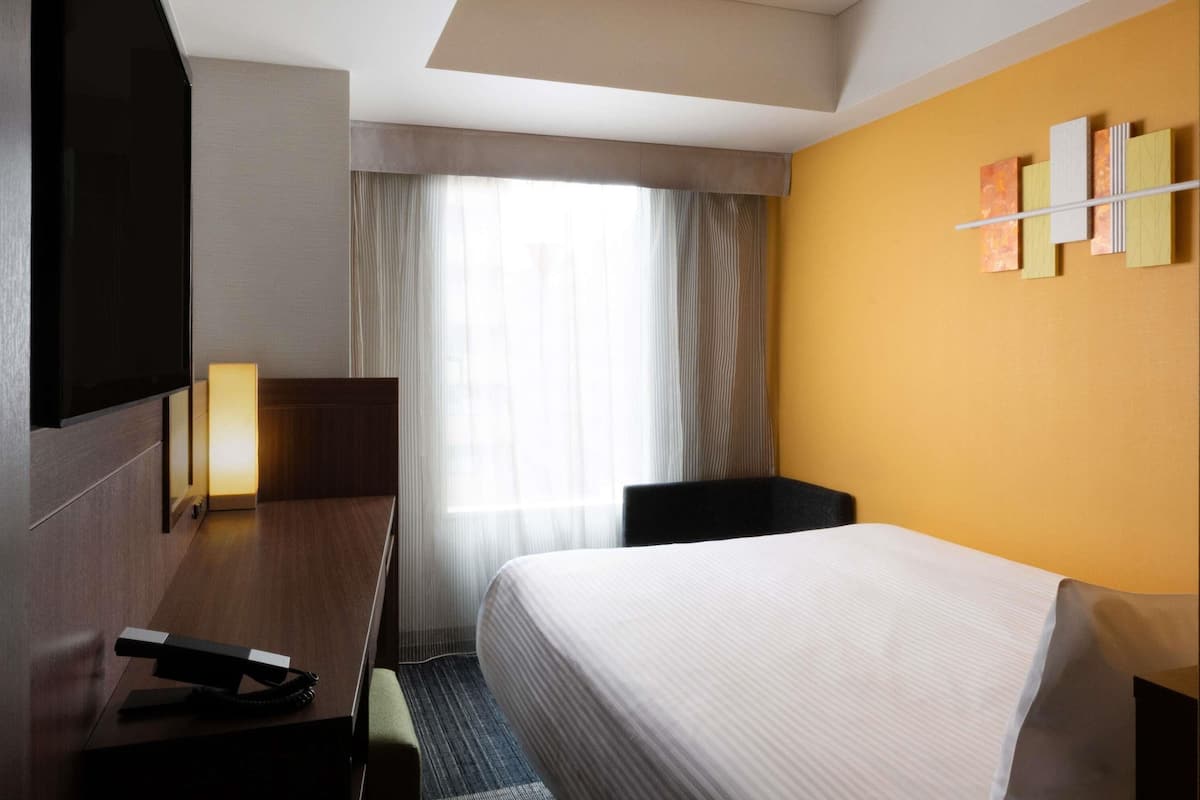 Een hotelkamer met een bed, een bureau met telefoon, een stoel, een televisie en een raam met gordijnen.