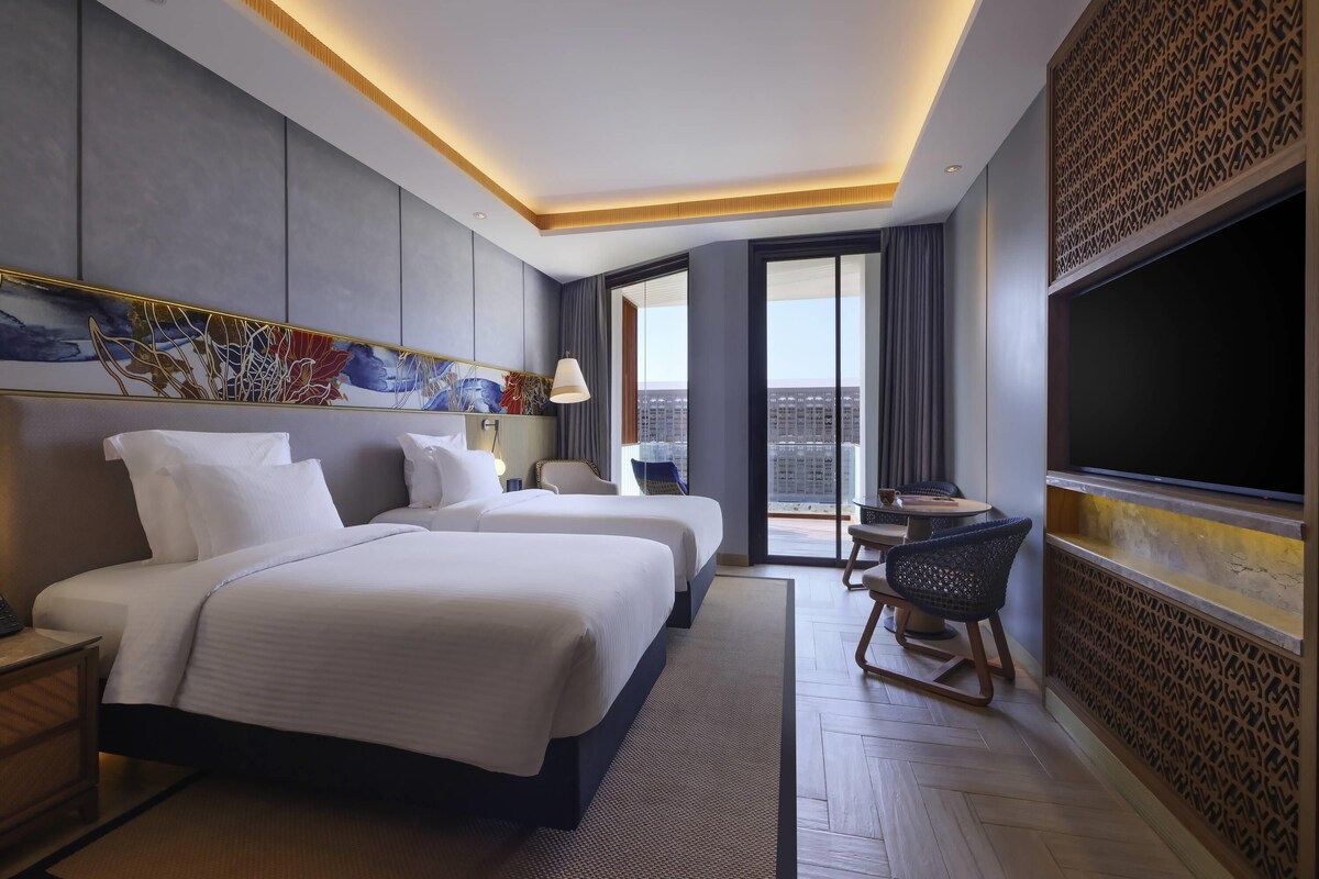 A hotel room with a large bed, a desk, a chair, a TV, and a view of the city.