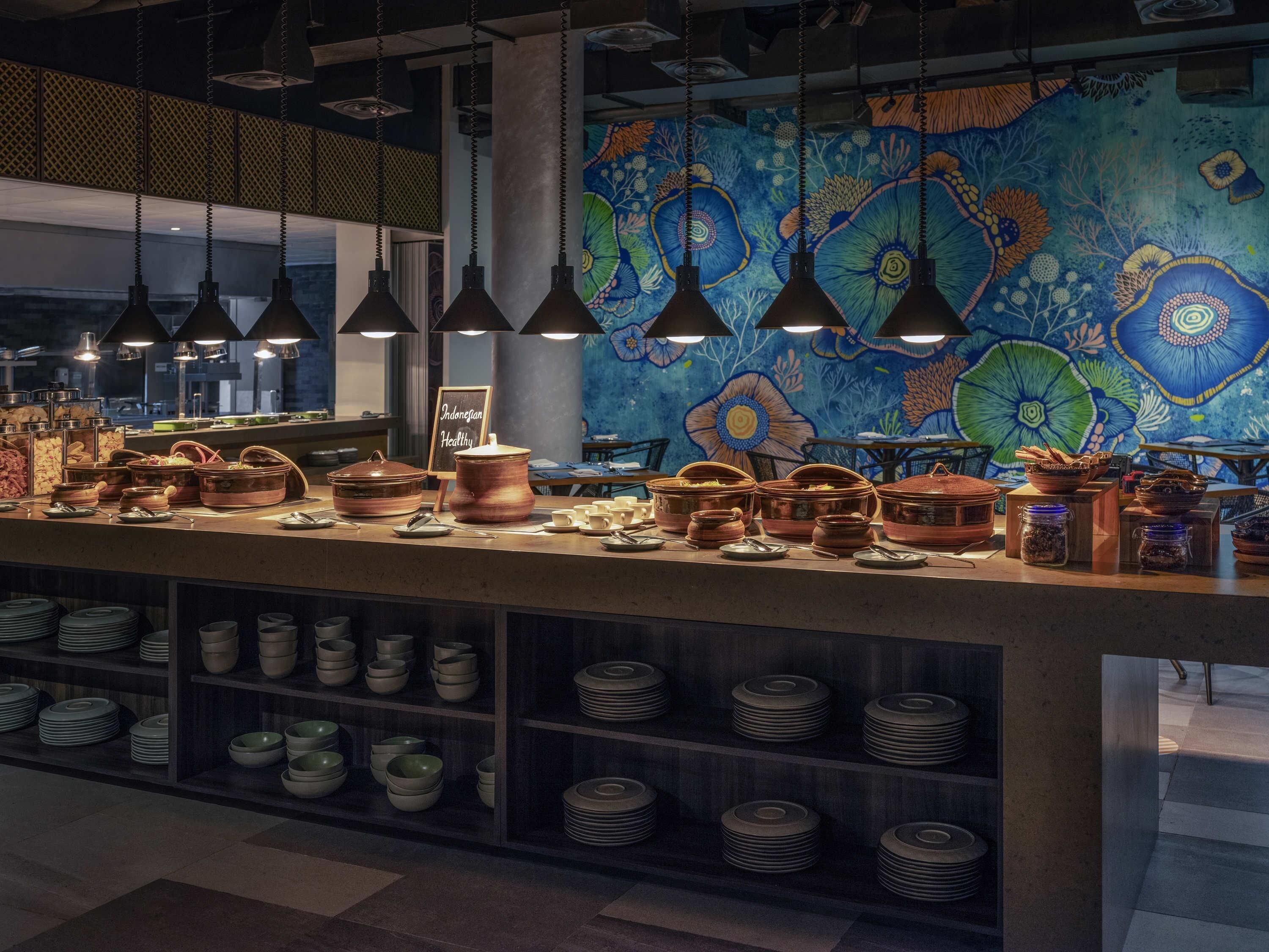 En restaurantdisk med forskellige retter og gryder, et vægmaleri og hængende pendellamper.