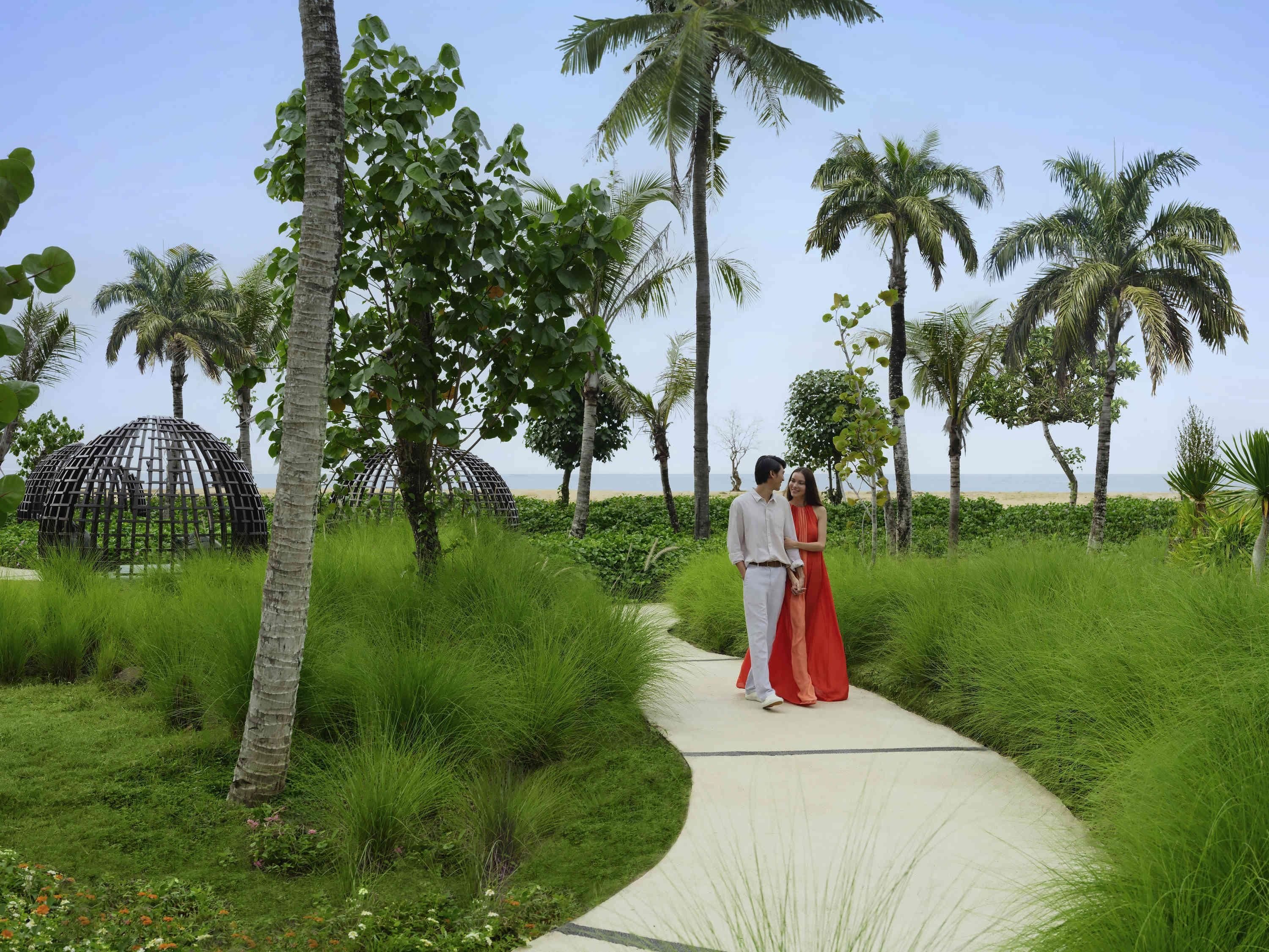 A pathway surrounded by lush greenery and palm trees, leading to a beach.