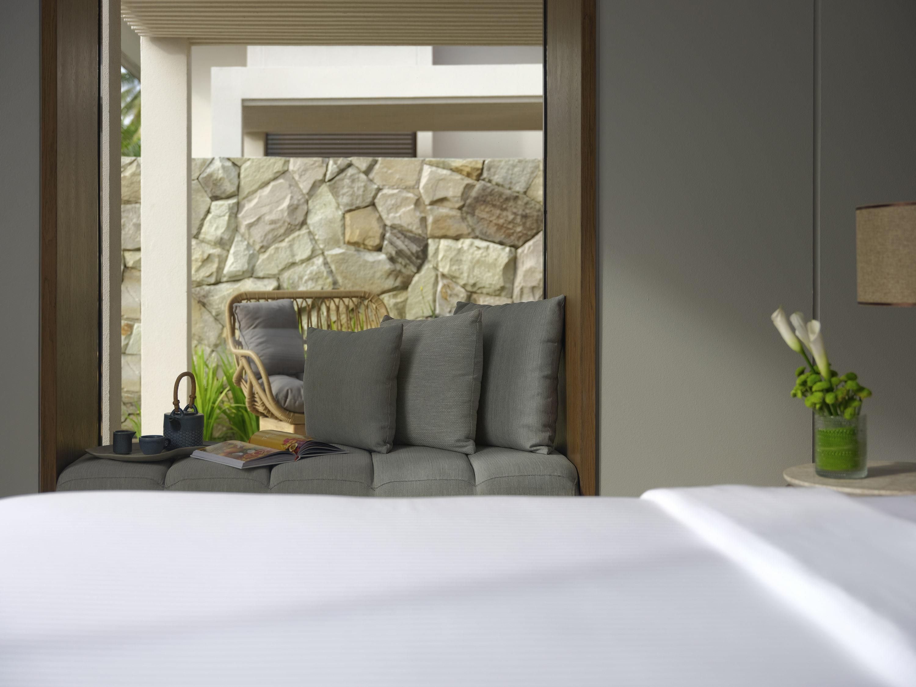 A modern hotel room with a stone wall, a grey sofa, a wicker chair, and a small table with a book and a teapot.