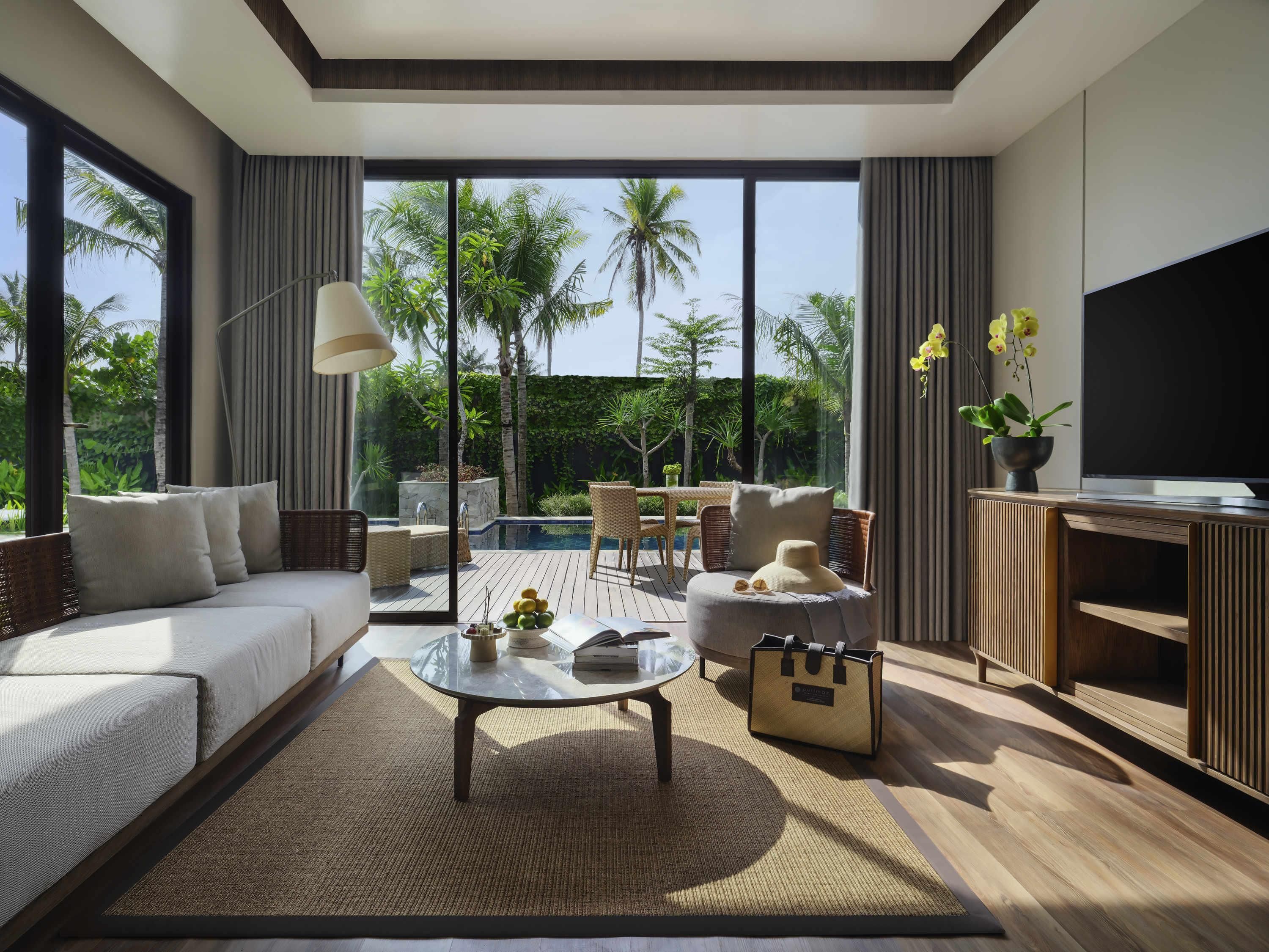 A modern living room with a sofa, coffee table, and TV, overlooking a pool and lush greenery.