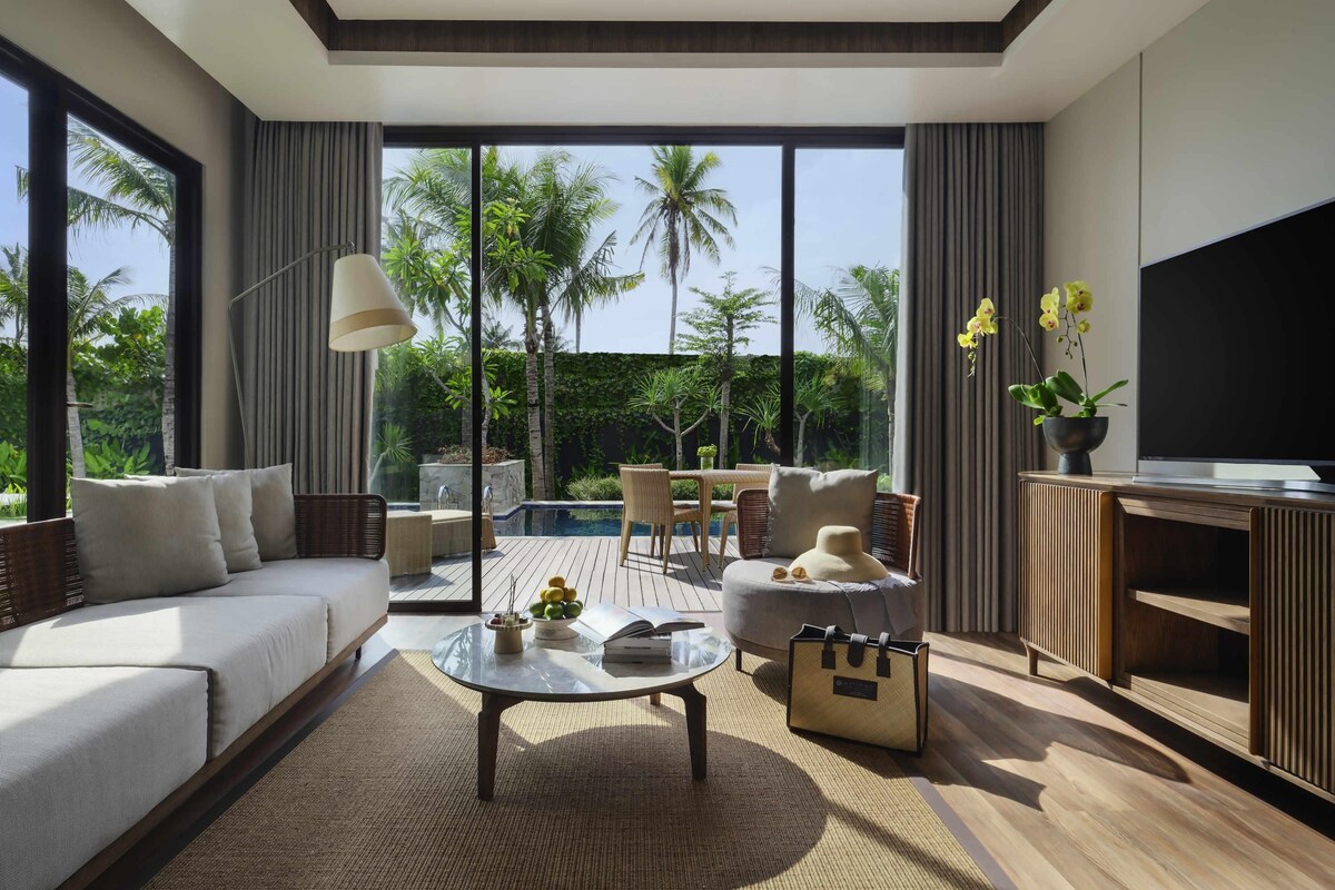 Un salon moderne avec un canapé, une table basse et un téléviseur, donnant sur une piscine et une végétation luxuriante.