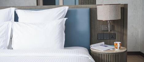A hotel room with a bed, pillows, a bedside table with a lamp, a mug, and a book.