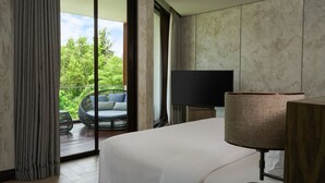 Une chambre d’hôtel avec un grand lit, une télévision, une lampe et un balcon doté d’un fauteuil en osier et offrant une vue sur la verdure.