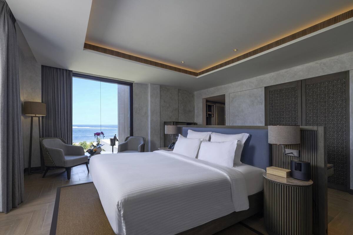 Une chambre d’hôtel moderne dotée d’un grand lit, d’un coin salon avec vue sur l’océan et d’une petite table avec une lampe.