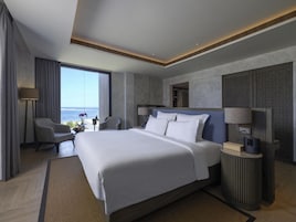 Una habitación de hotel moderna con una cama grande, una zona de estar con vistas al océano y una mesita con lámpara.