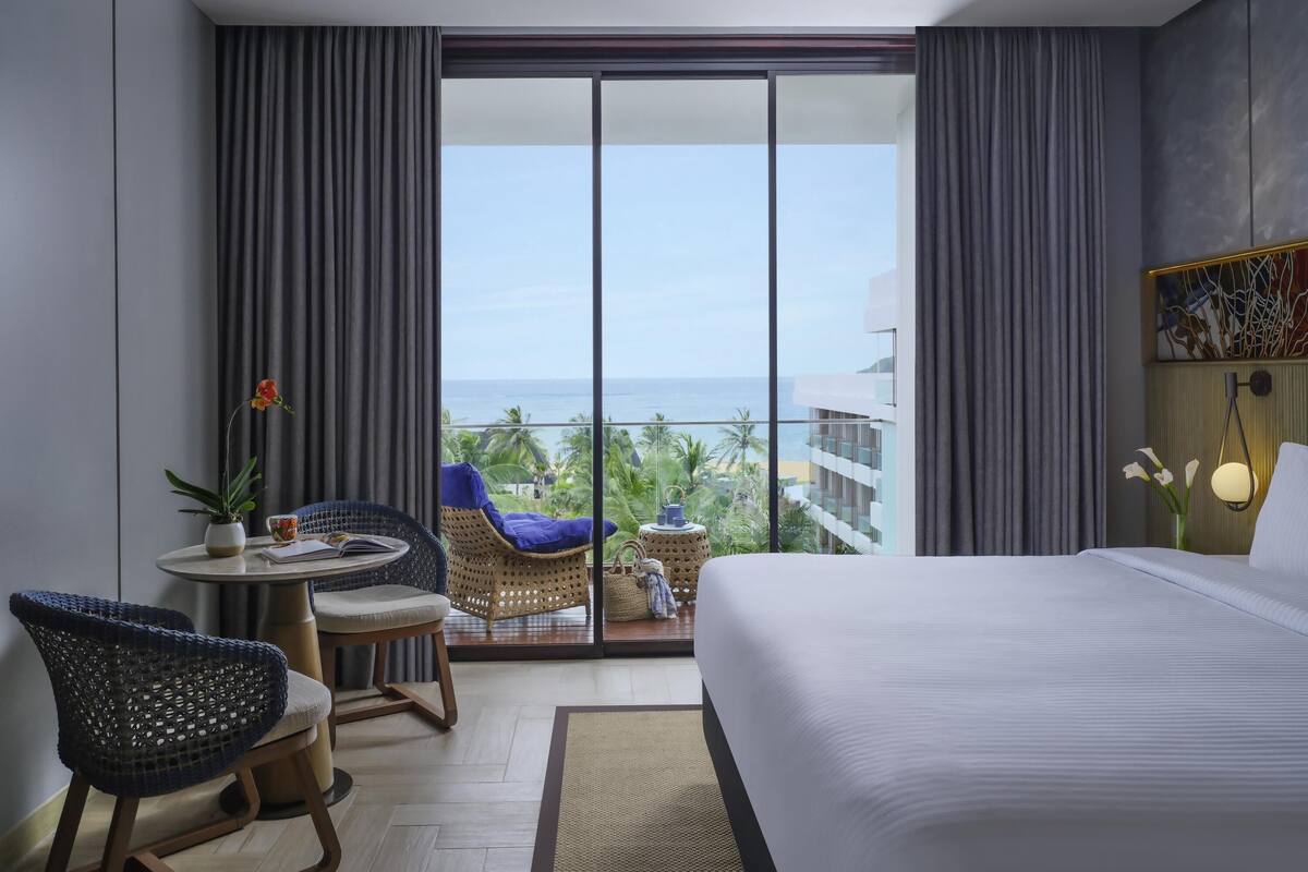 Une chambre d’hôtel avec un lit, une table ronde entourée de chaises, une vue sur l’océan et un balcon agrémenté d’un fauteuil en osier.