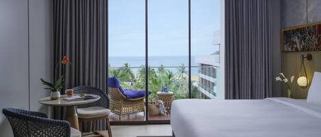 A hotel room with a bed, a round table with chairs, a view of the ocean, and a balcony with a wicker chair.