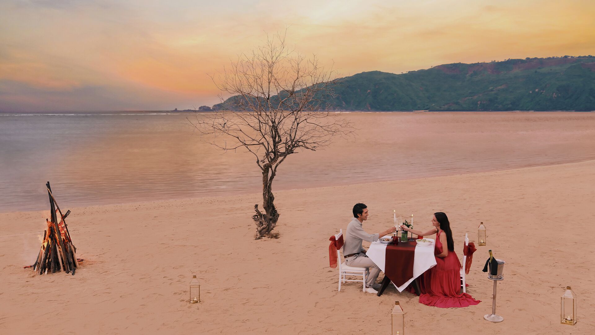 A couple dining on a beach with a table set for two, a single tree, and a campfire in the background.