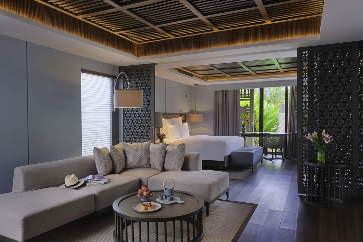 Une chambre d’hôtel moderne, dotée d’un canapé, d’une table basse ronde, d’un lit et d’un balcon offrant une vue sur la verdure.