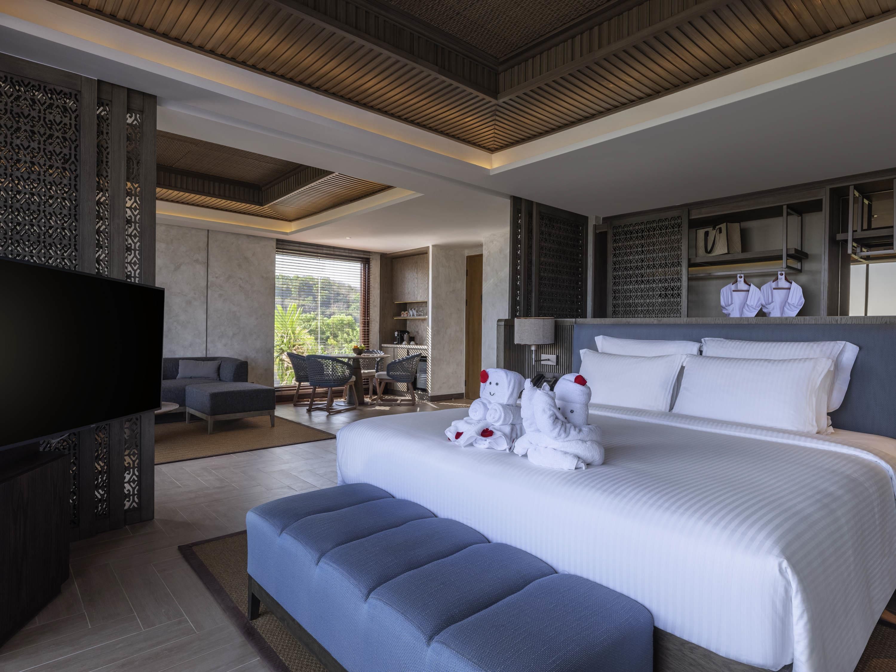 A modern hotel room with a large bed, a flat-screen TV, a seating area with chairs, and a view of the outdoors.