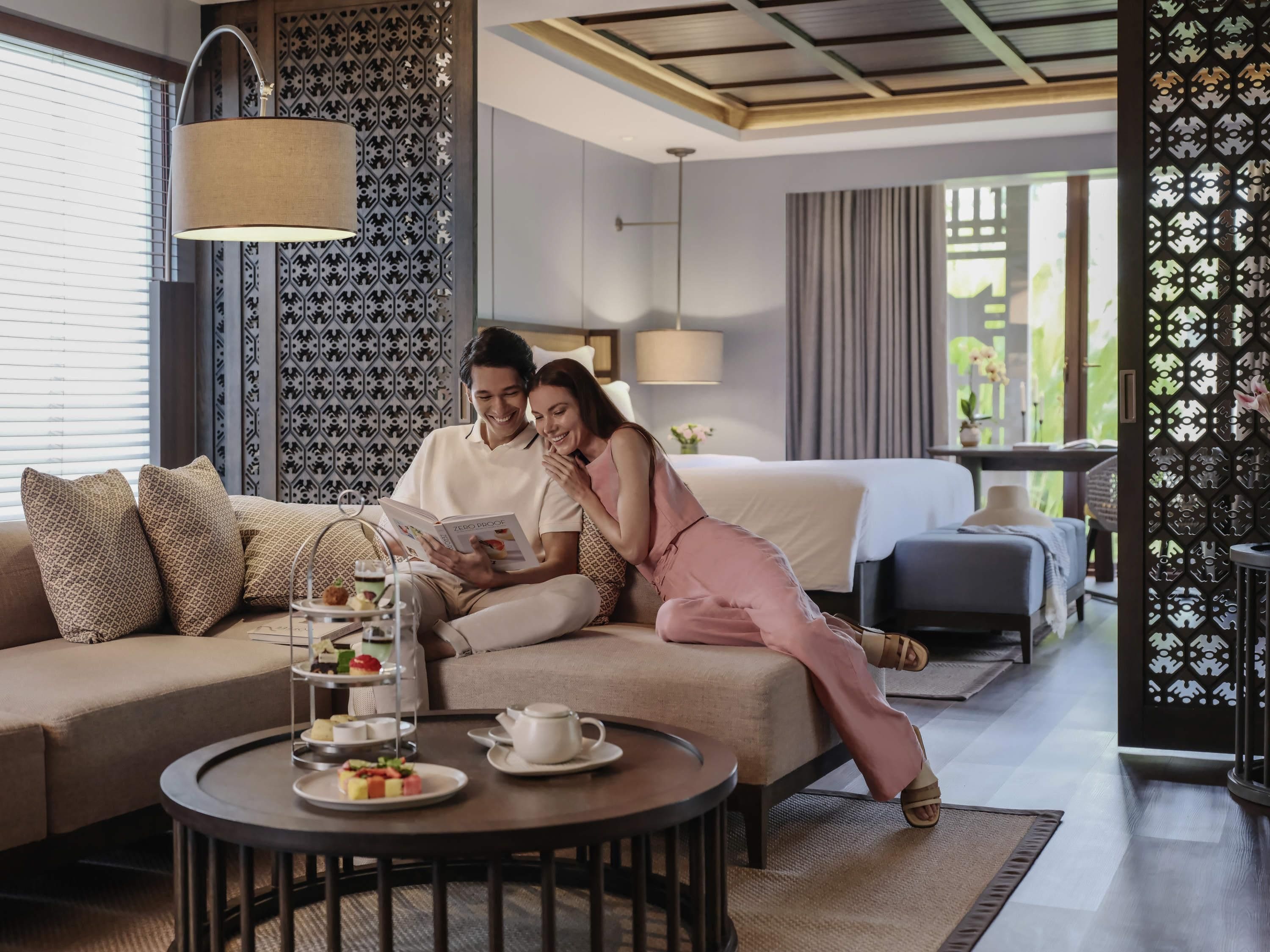 Une chambre d’hôtel moderne avec un canapé, une table basse sur laquelle sont disposées des pâtisseries, un lit et un élément de séparation décoratif.