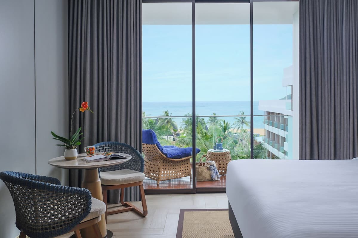 A hotel room with a balcony, a bed, a small table with a plant, two chairs, and a view of the ocean.